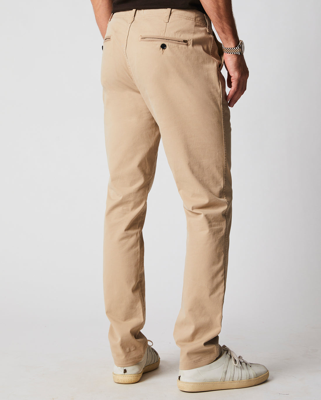 A man is standing facing away, wearing beige chinos, a black shirt, a silver watch, and white sneakers against a plain light background.