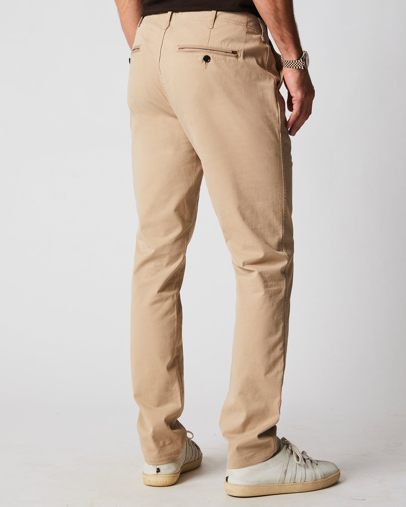 A man is standing facing away, wearing beige chinos, a black shirt, a silver watch, and white sneakers against a plain light background.