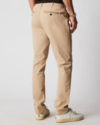 A man is standing facing away, wearing beige chinos, a black shirt, a silver watch, and white sneakers against a plain light background.