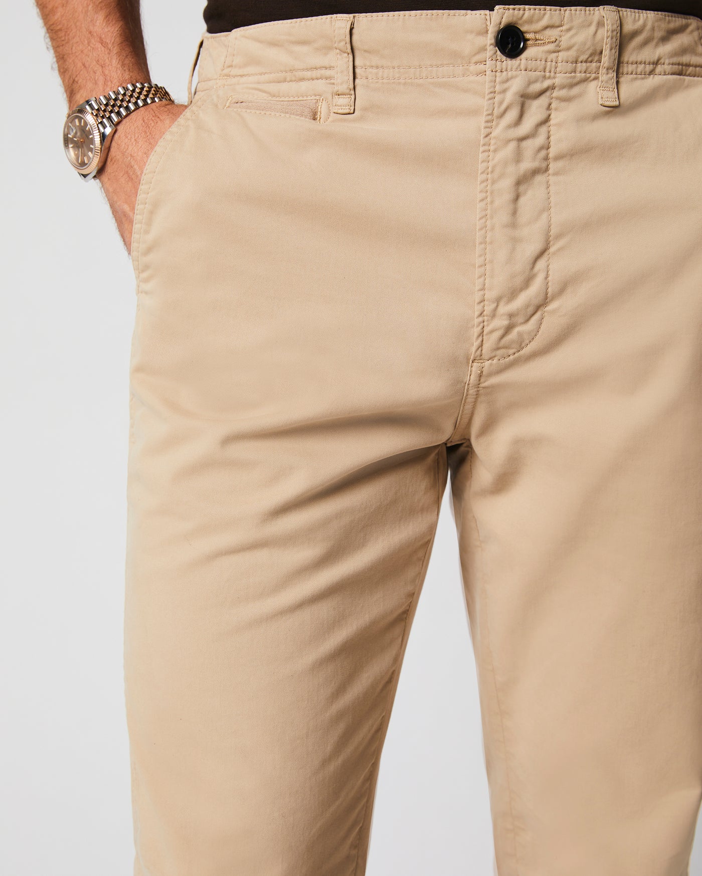 A person wearing beige khaki pants and a watch with a metal band, with one hand in a pocket, against a plain background.