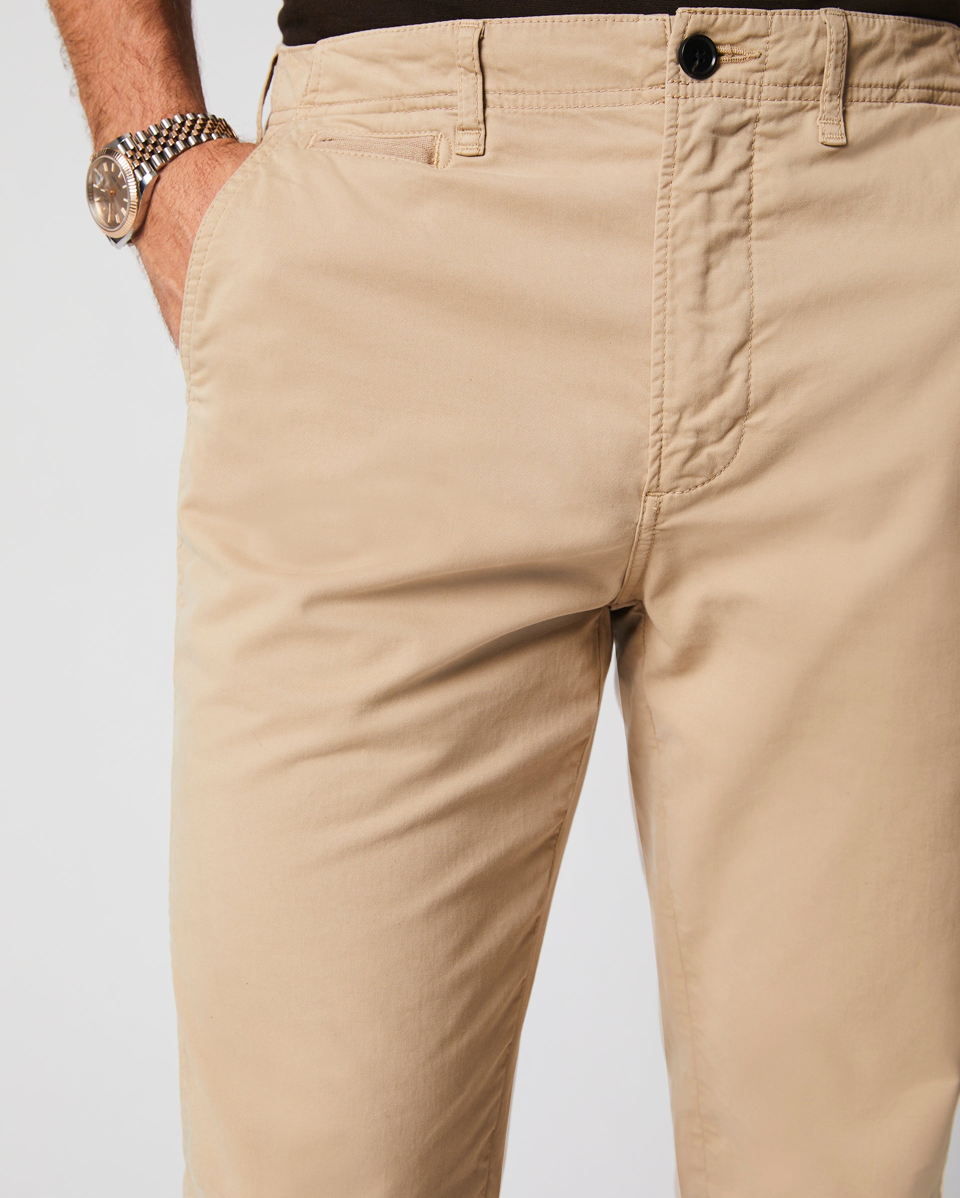 A person wearing beige khaki pants and a watch with a metal band, with one hand in a pocket, against a plain background.