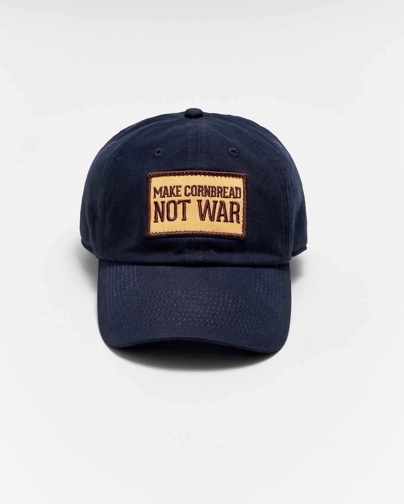 A navy blue baseball cap with a tan patch on the front that reads MAKE CORNBREAD NOT WAR in bold capital letters. The hat is photographed against a plain white background.