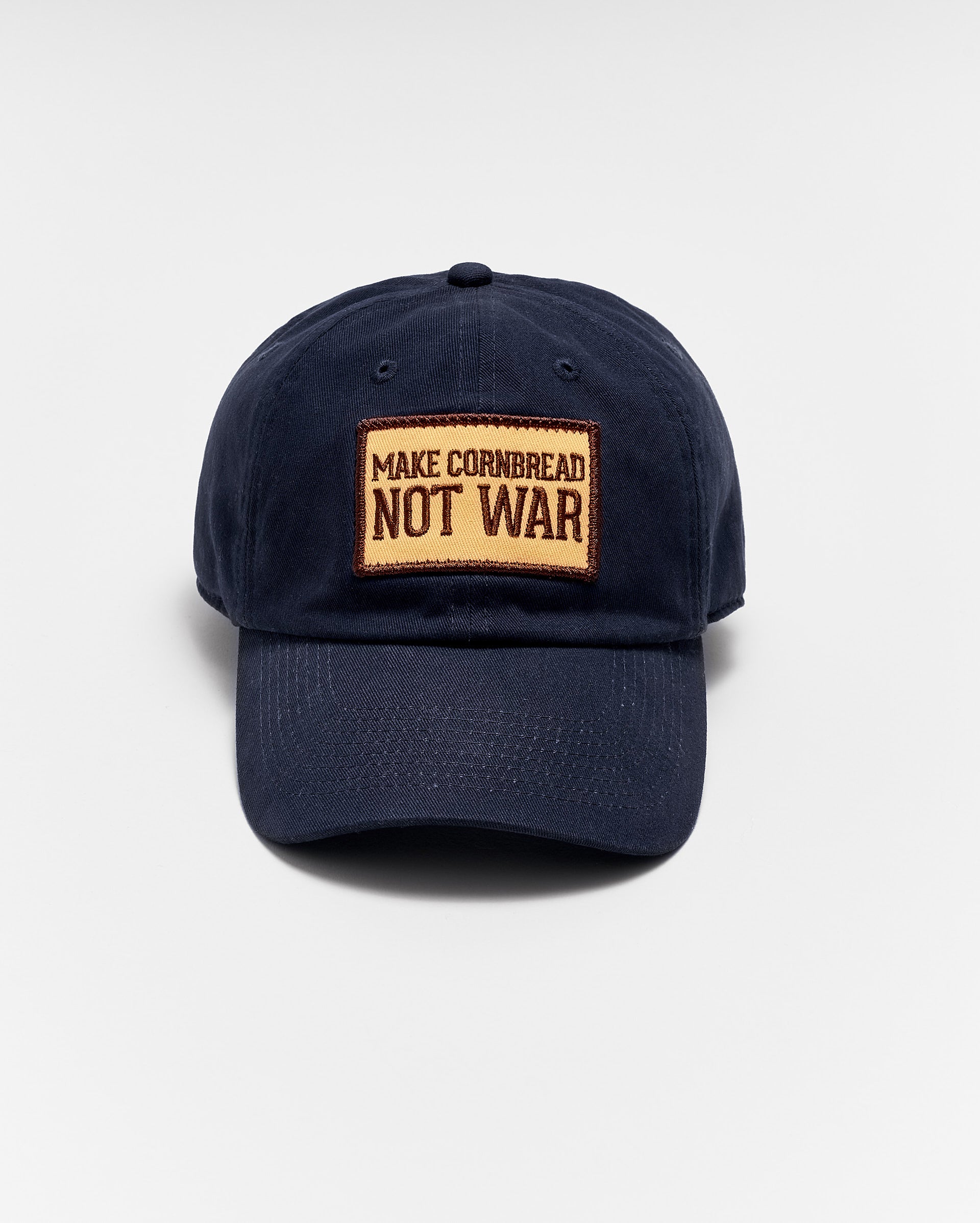 A navy blue baseball cap with a tan patch on the front that reads MAKE CORNBREAD NOT WAR in bold capital letters. The hat is photographed against a plain white background.