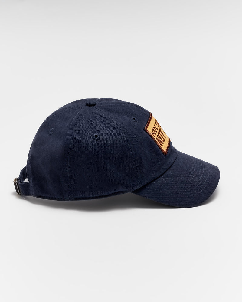 A navy blue baseball cap with a brown rectangular patch on the front, viewed from the side against a plain white background. The cap has an adjustable strap at the back. Alternate