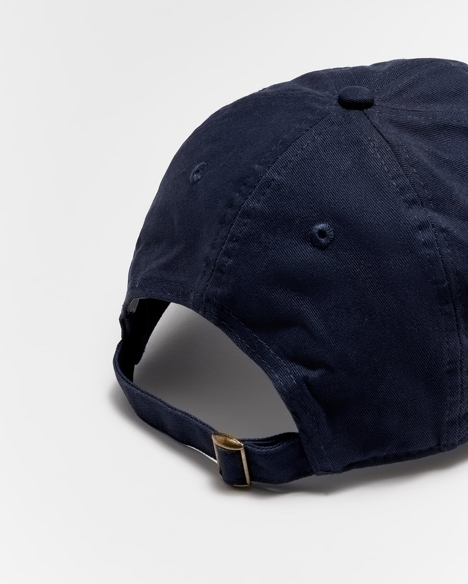 A navy blue baseball cap shown from the back, featuring a fabric strap with a brass buckle for adjusting the fit. The cap is made of cotton and photographed on a plain white background.