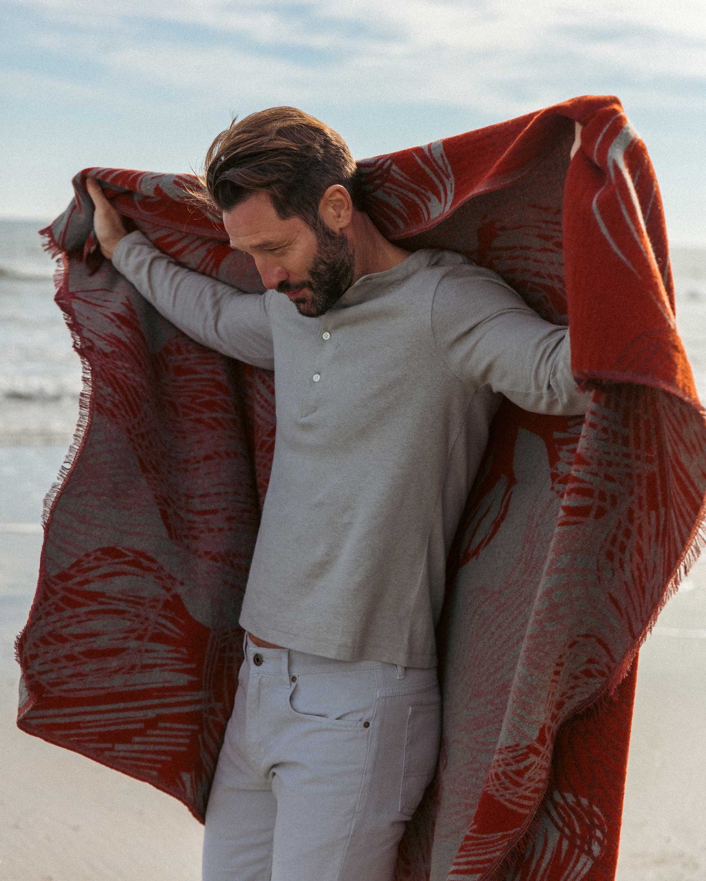 A man with a beard stands on a beach, looking down and smiling, while draping a large red and gray patterned blanket over his shoulders. He wears a light gray long-sleeve shirt and light-colored pants.